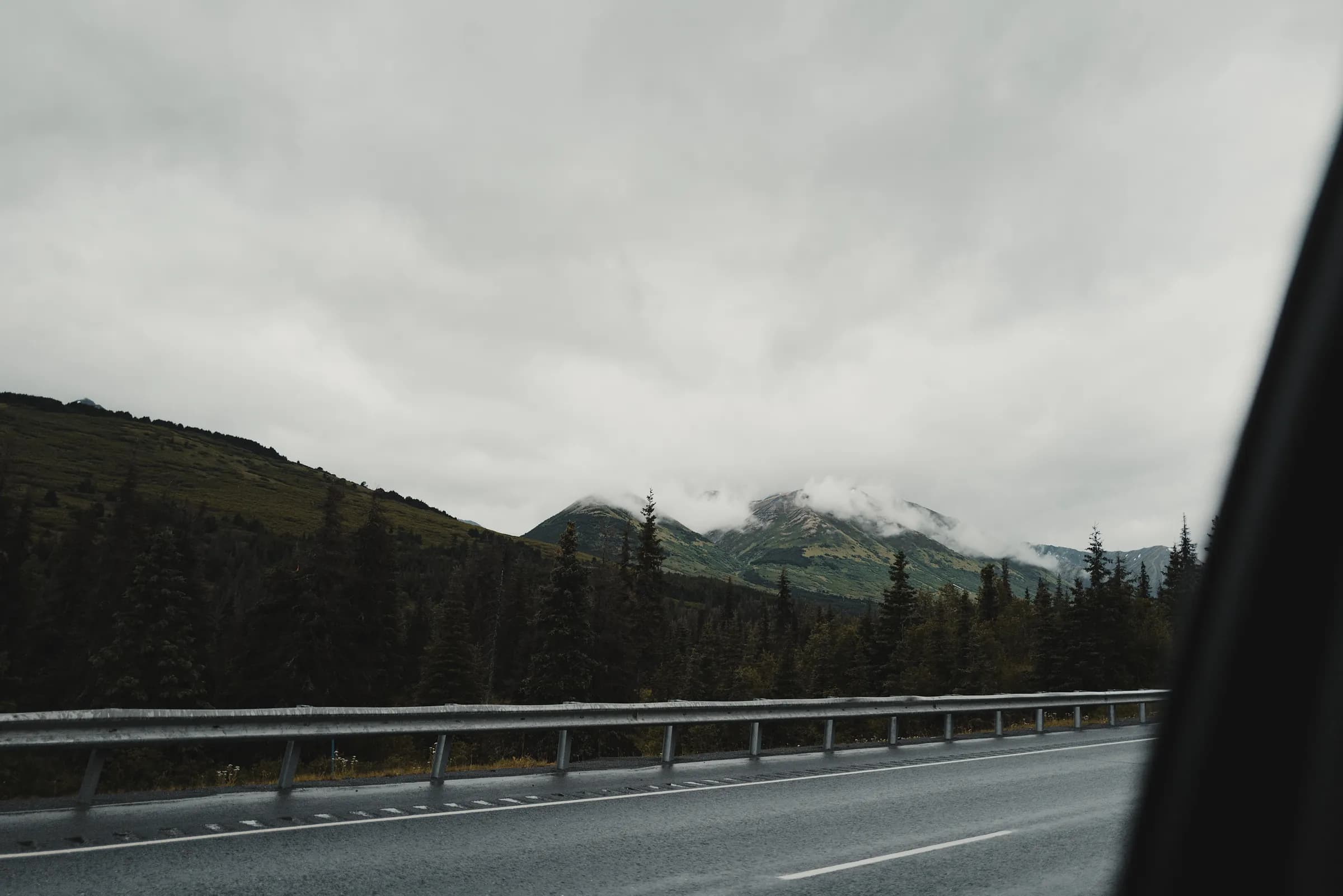 A wet highway and guardrail beneath cloud-covered Alaskan mountains, seen from a moving vehicle