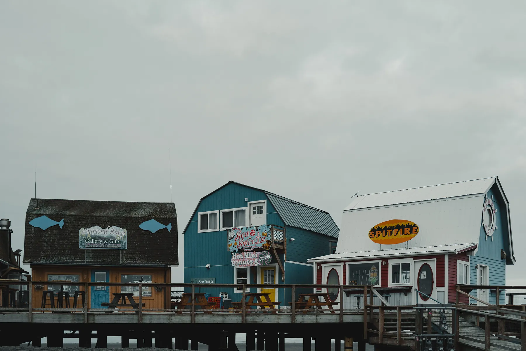 Painted wooden shopfronts on stilts along the Homer Spit boardwalk under a grey sky