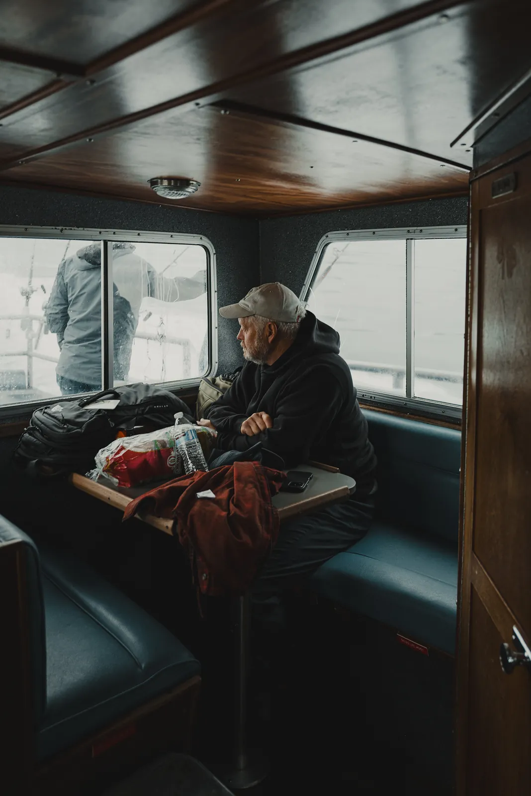 A man in a cap sitting at a boat cabin table looking out the window at grey water