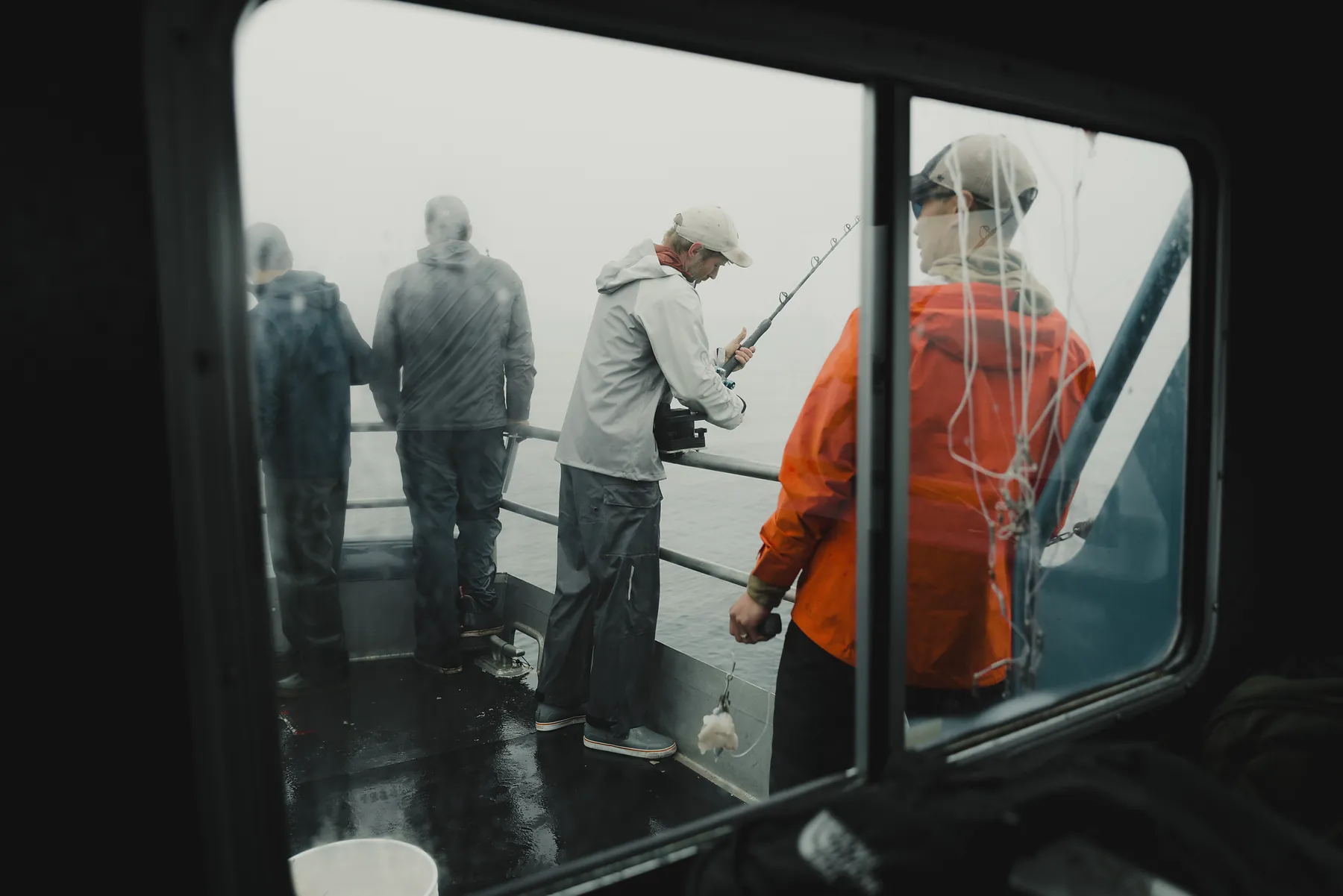 Anglers in raingear standing along a boat rail, seen through a wheelhouse window