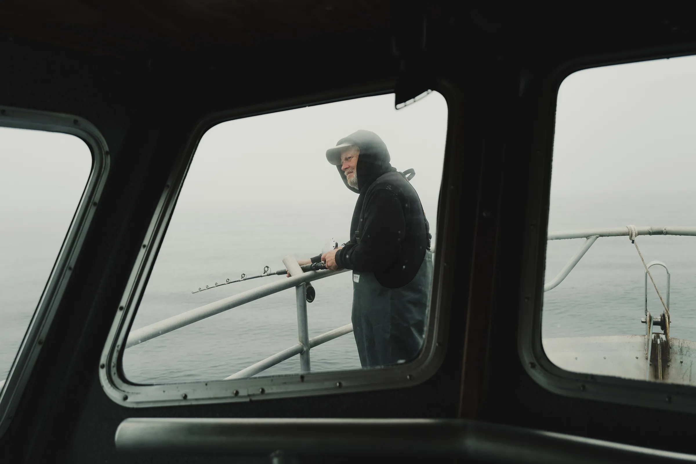 A hooded angler working a rod at the boat rail, framed through the wheelhouse windows in fog