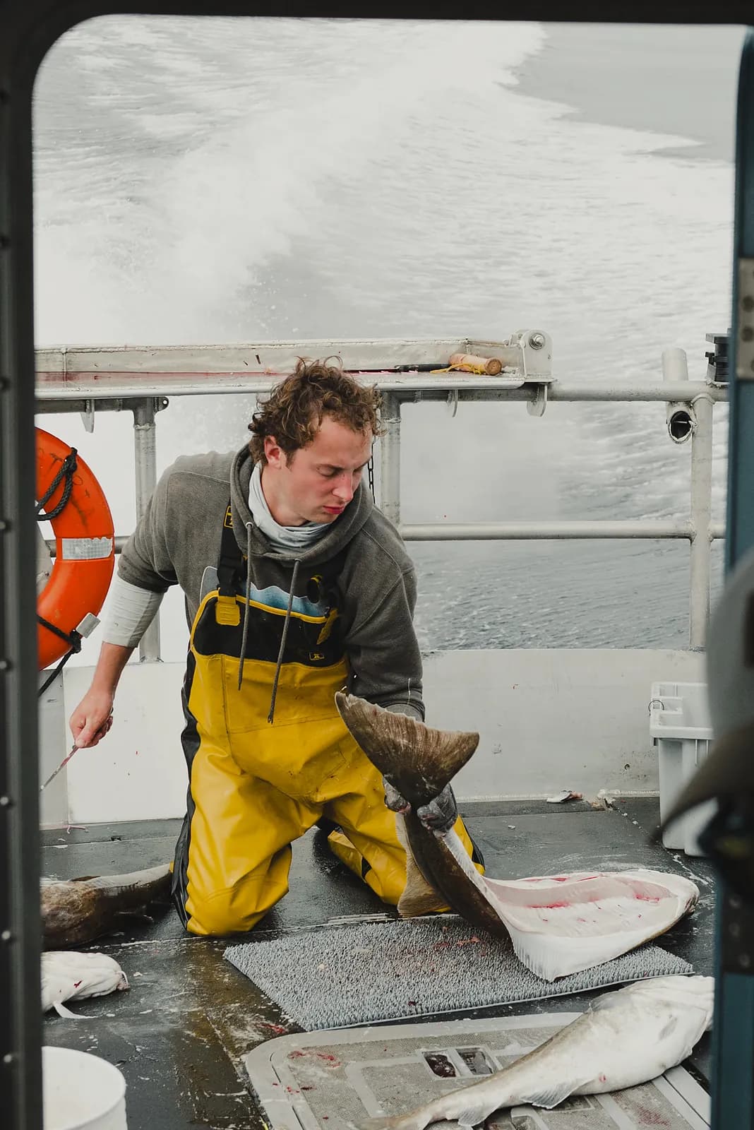 A deckhand in yellow waders filleting fish on the open deck of a moving boat