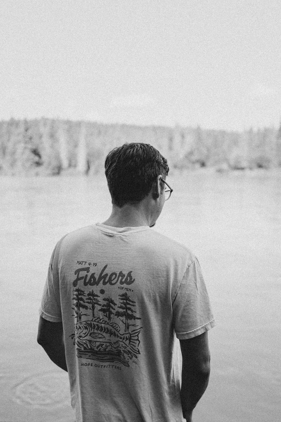 Black and white photograph of a man from behind wearing a shirt reading Fishers of Men, facing a wide river
