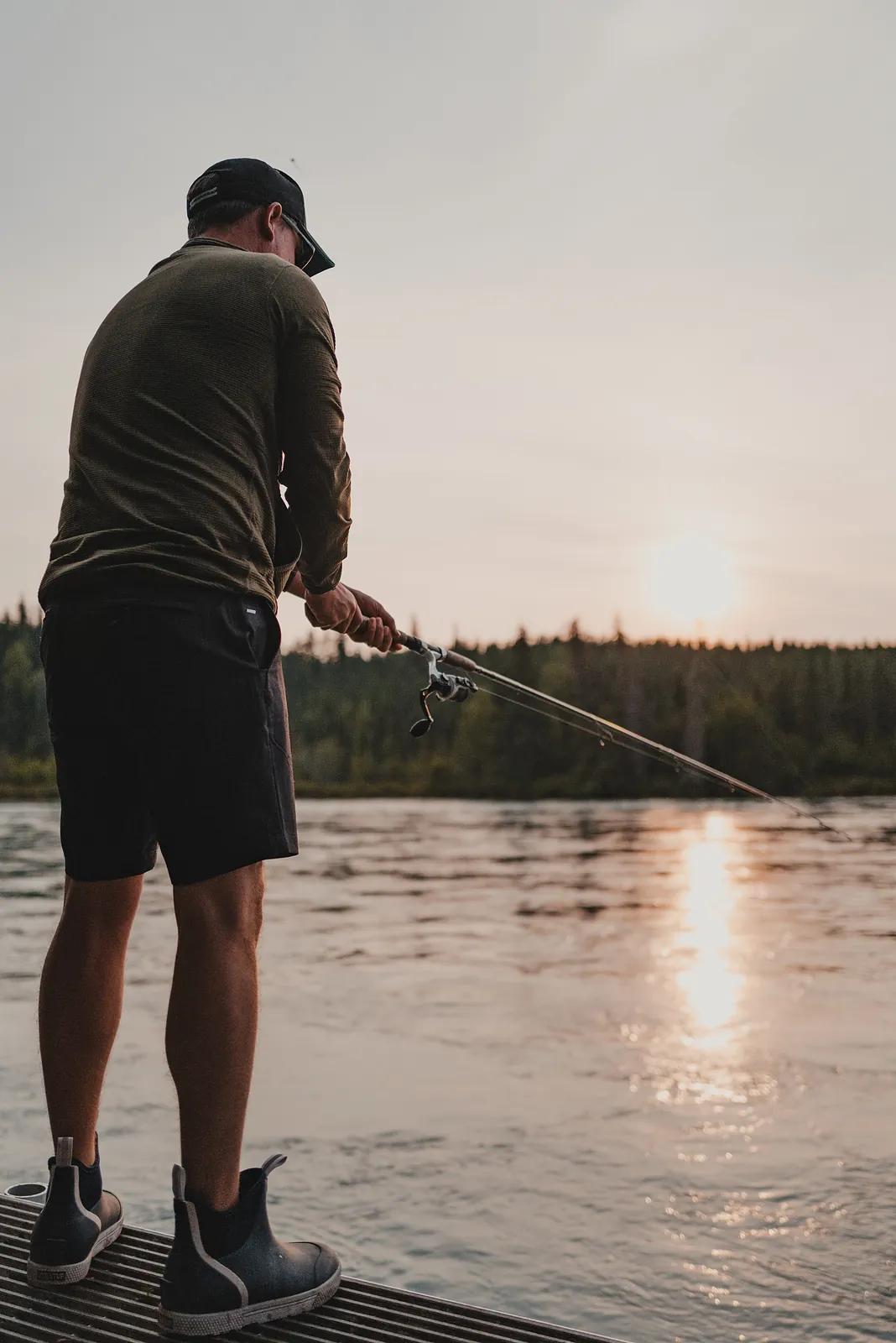 A man casting from a dock into a river carrying a long gold reflection of low sun