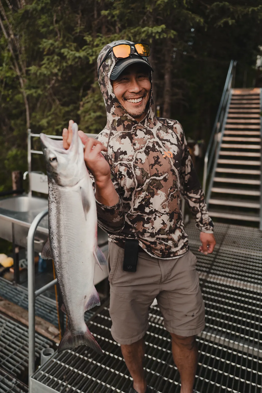 A smiling man in camouflage holding up a large silver salmon on a dock stairway