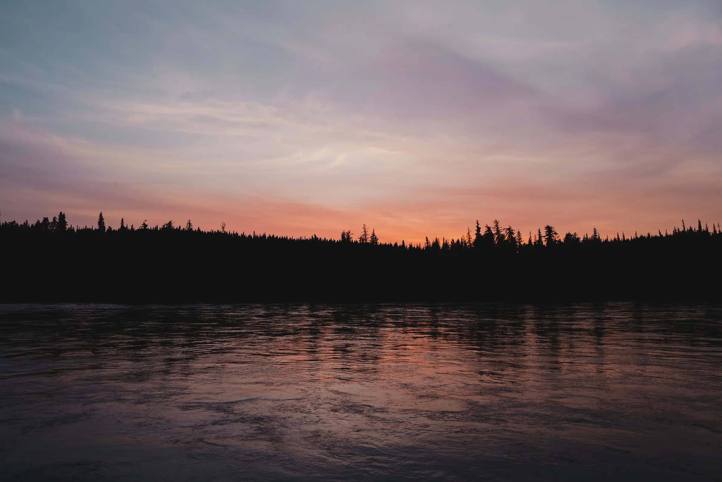A pink and orange sunset spread along a black spruce treeline above a still river