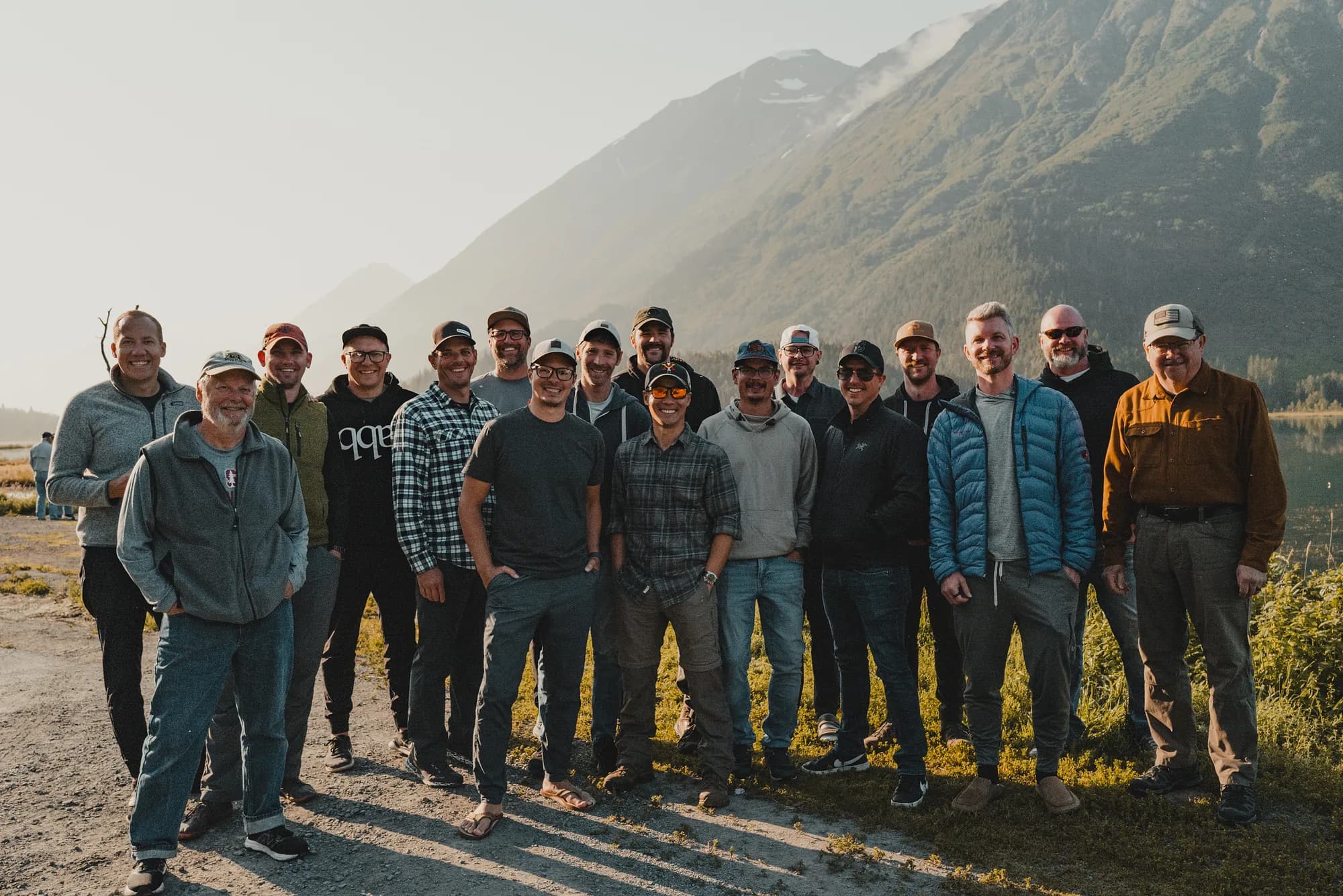 A large group of men standing in a line on a gravel pullout below Alaskan mountains