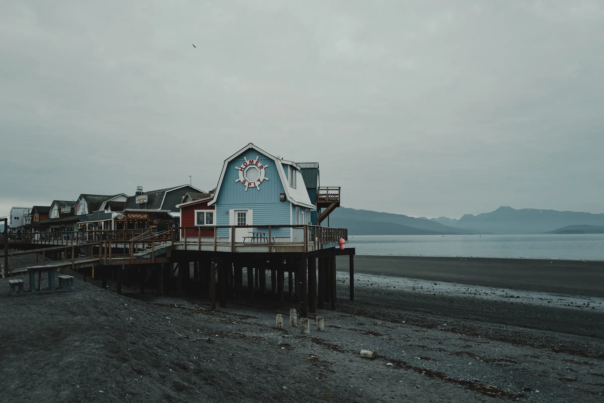 A blue building on pilings at the end of the Homer Spit above a dark gravel beach, mountains beyond