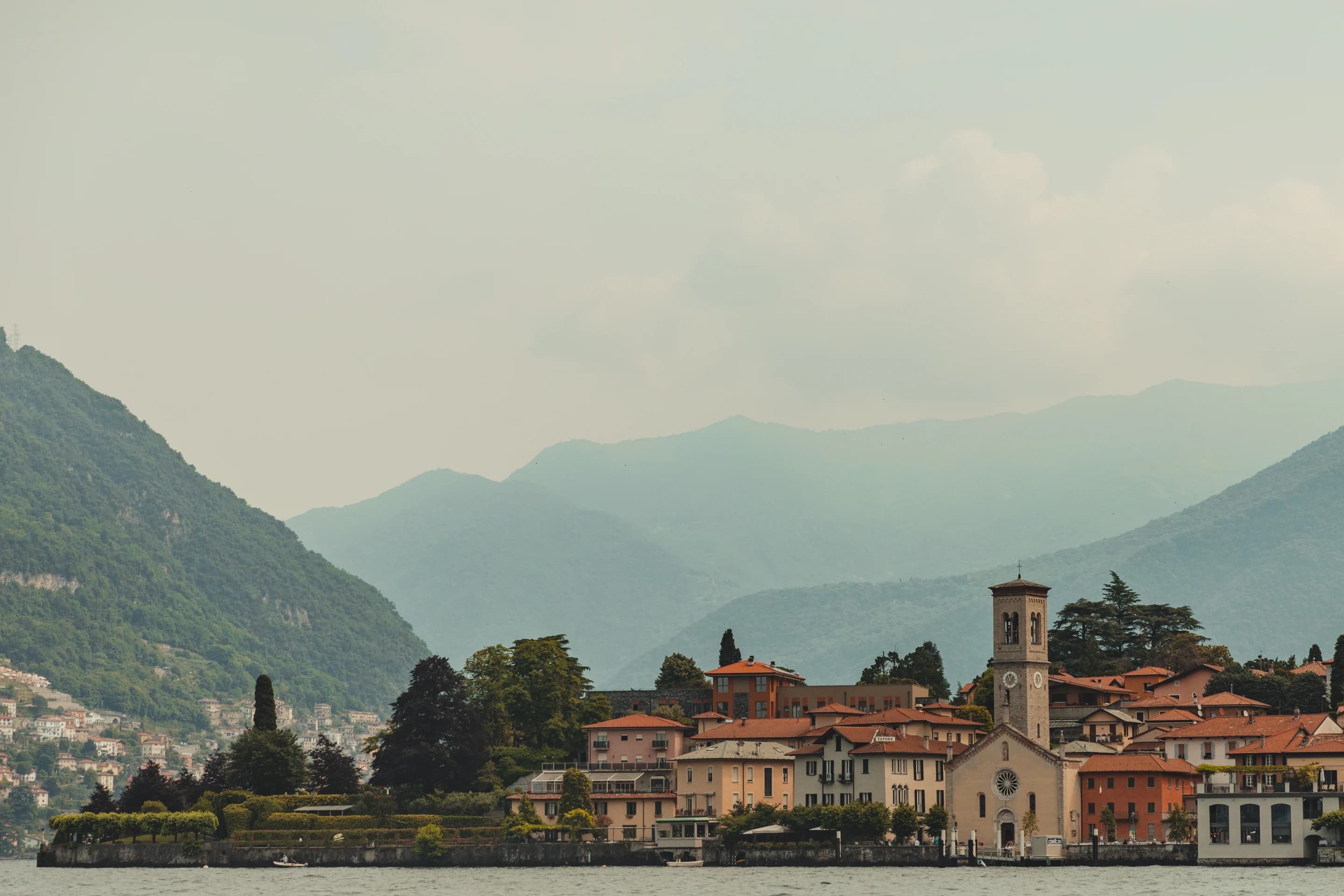 A village and church on the shore of Lake Como beneath hazy mountains