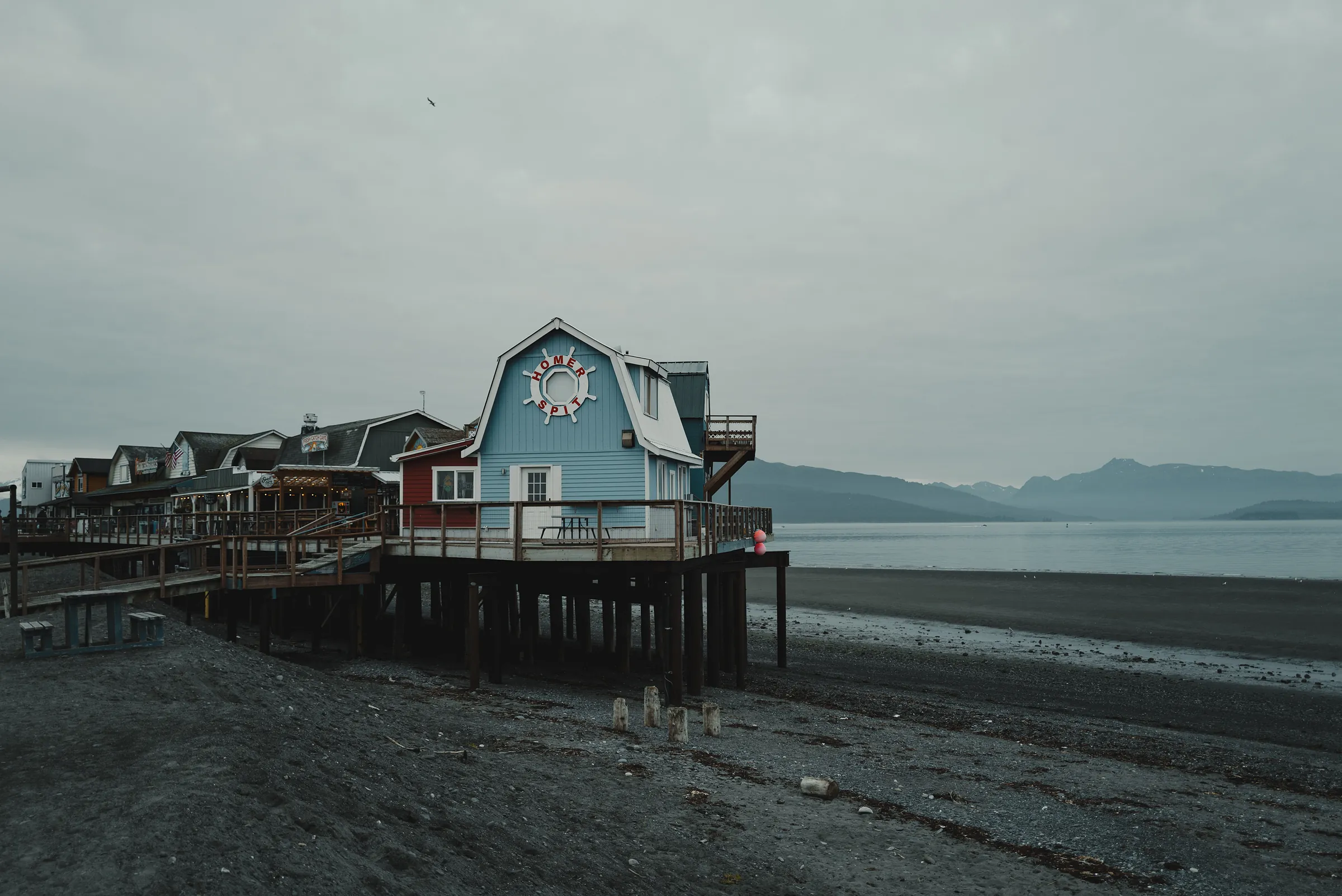 A blue building on pilings at the end of the Homer Spit above a dark gravel beach